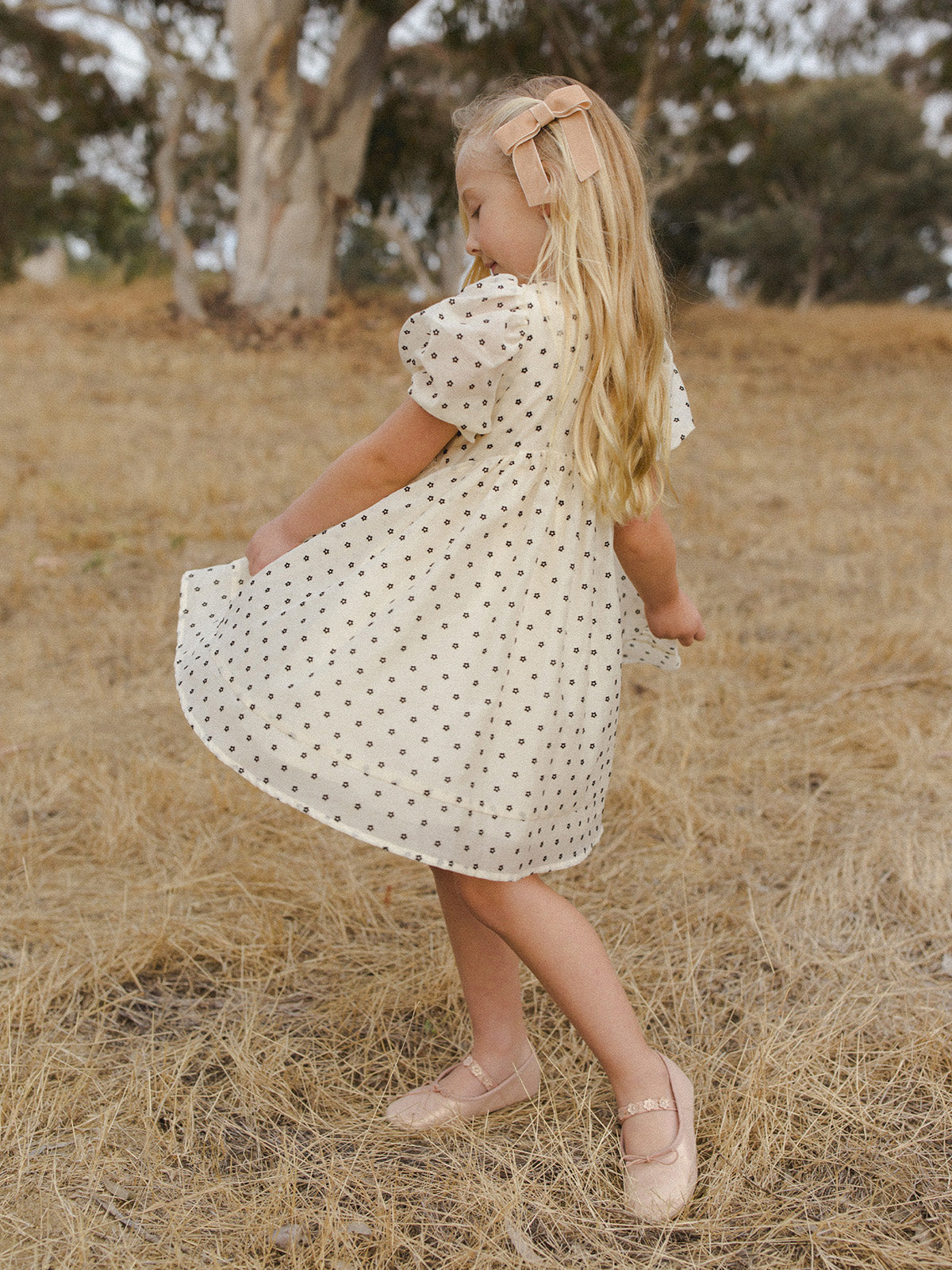 Young girl in a polka dot dress twirls in a golden field, wearing ballet flats and a hair bow.