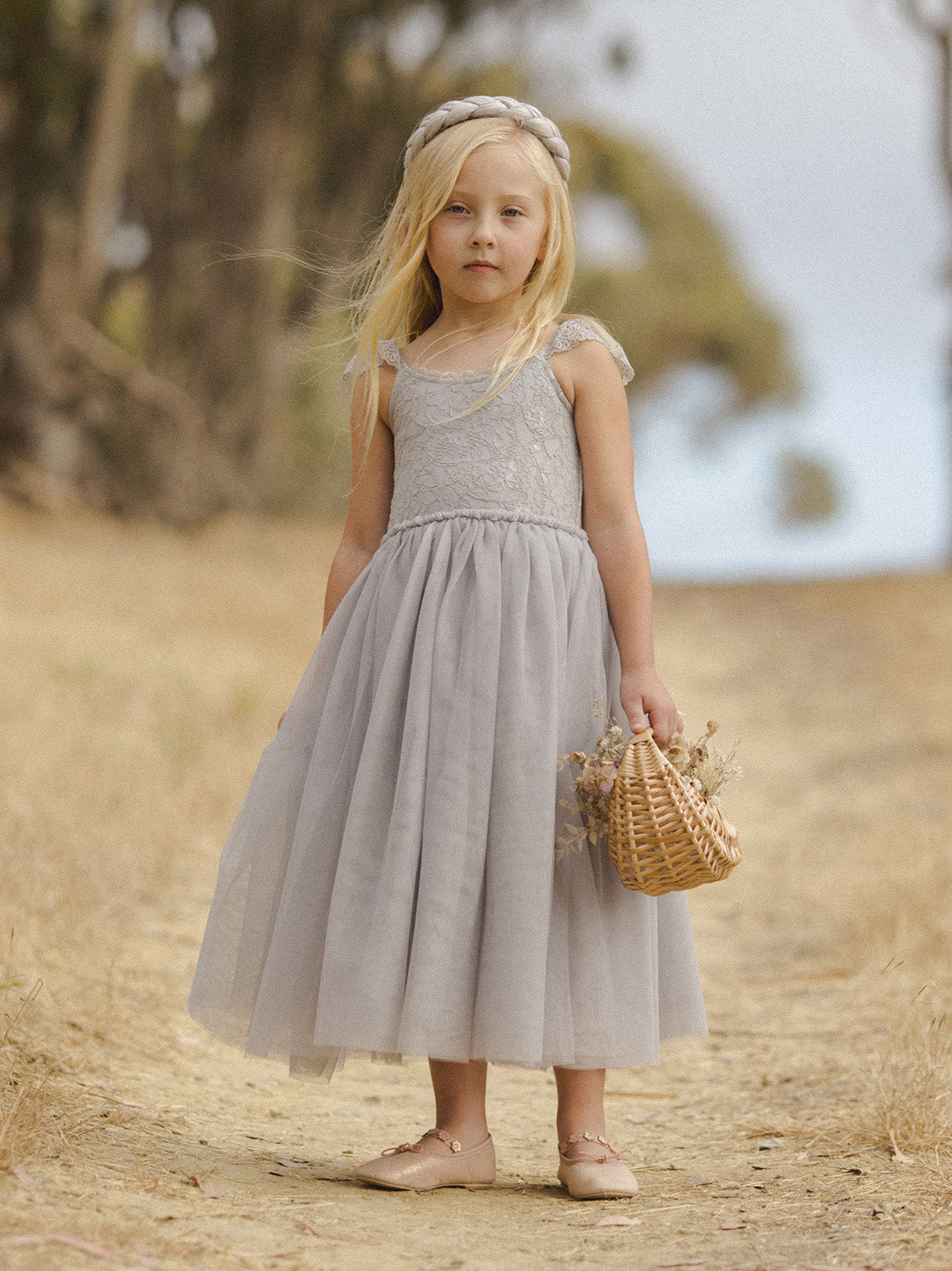 Girl in a gray dress with a basket stands on a dirt path, surrounded by blurred greenery.