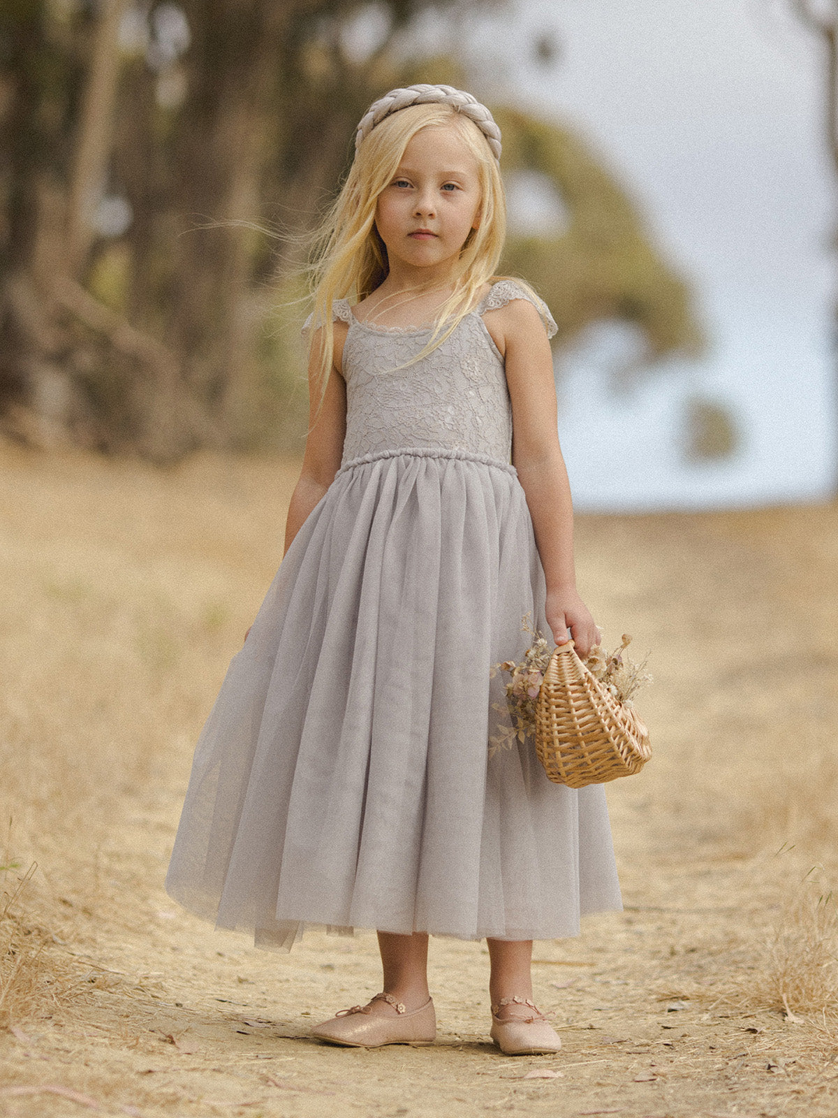 Girl in a gray dress with a basket stands on a dirt path, surrounded by blurred greenery.