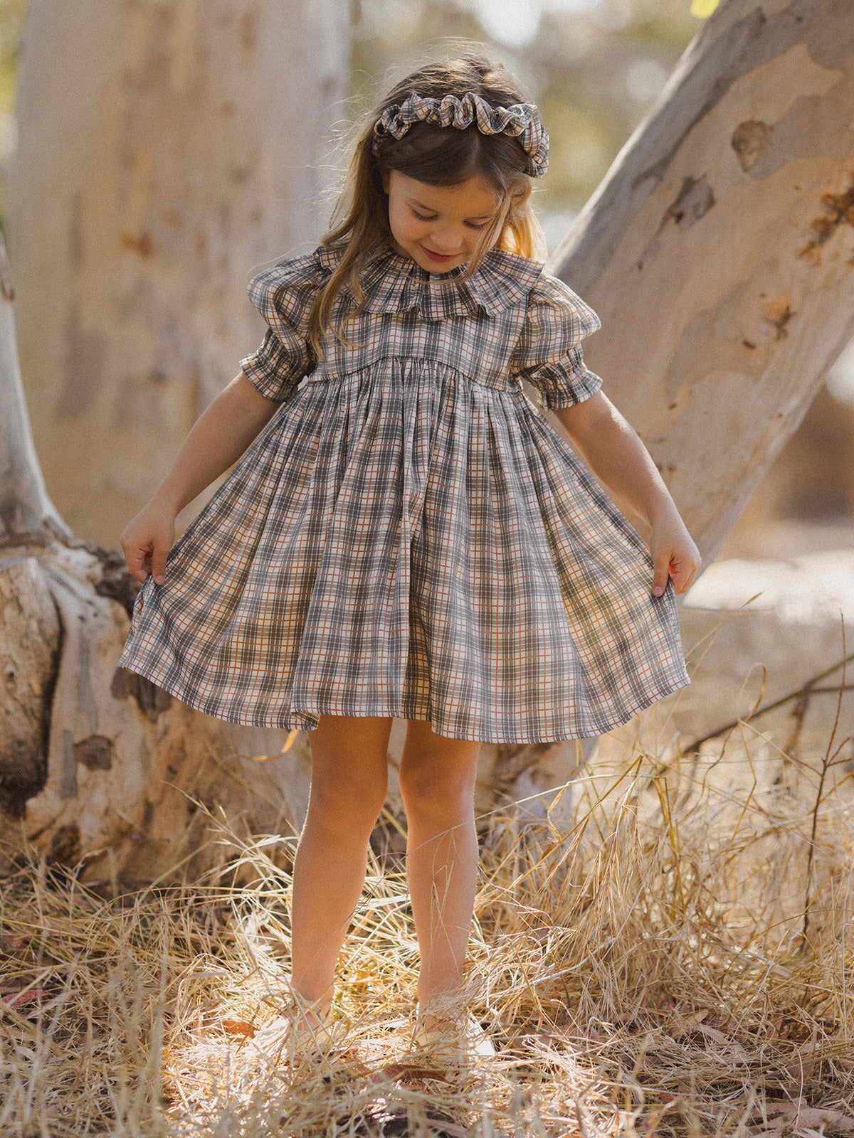 Girl in a plaid dress with puffed sleeves, smiling and playing outdoors near a tree.