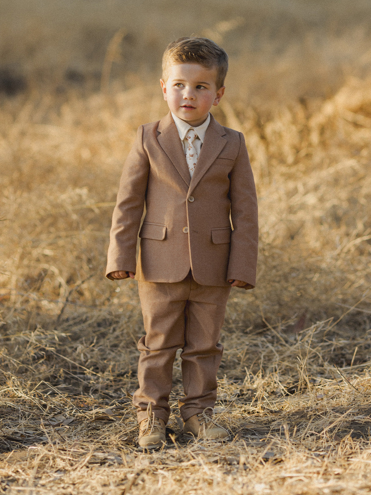 Young boy in a brown suit stands in a field of dry grass, looking thoughtfully ahead.