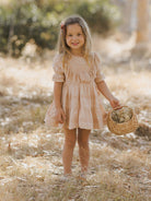 Smiling girl in a light dress holding a small basket in a sunlit natural setting.