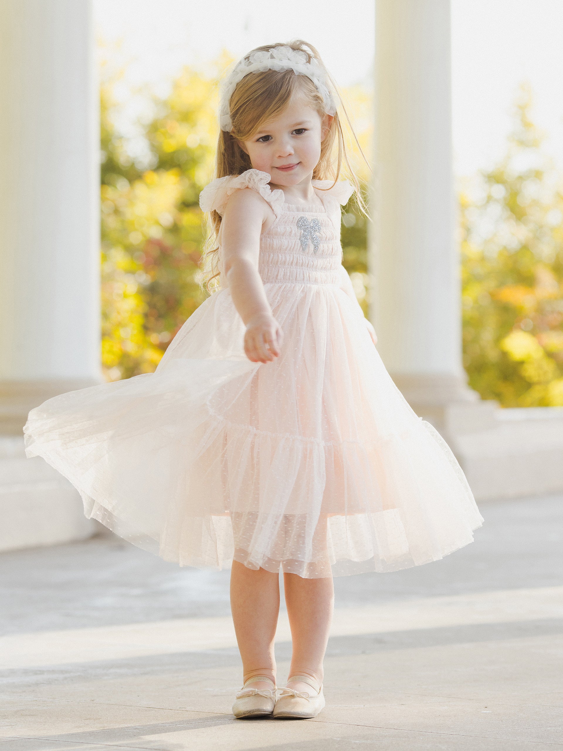 Young girl in a light pink dress twirling, smiling, with a headband, in an outdoor setting with soft sunlight.