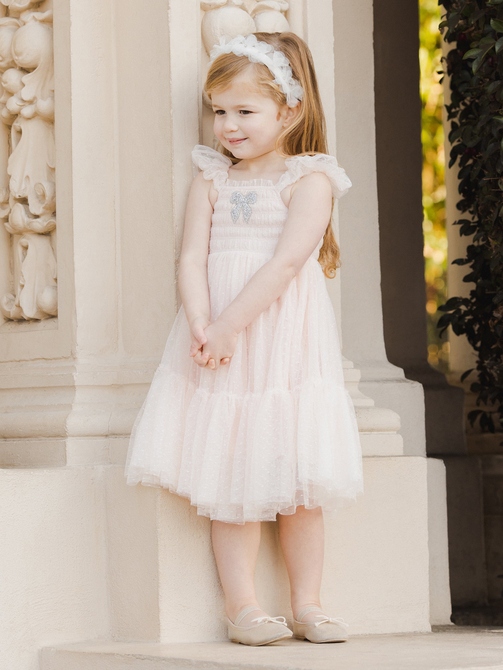 Young girl wearing a light pink dress, smiling next to elegant architecture, with a floral headband and ballet flats.