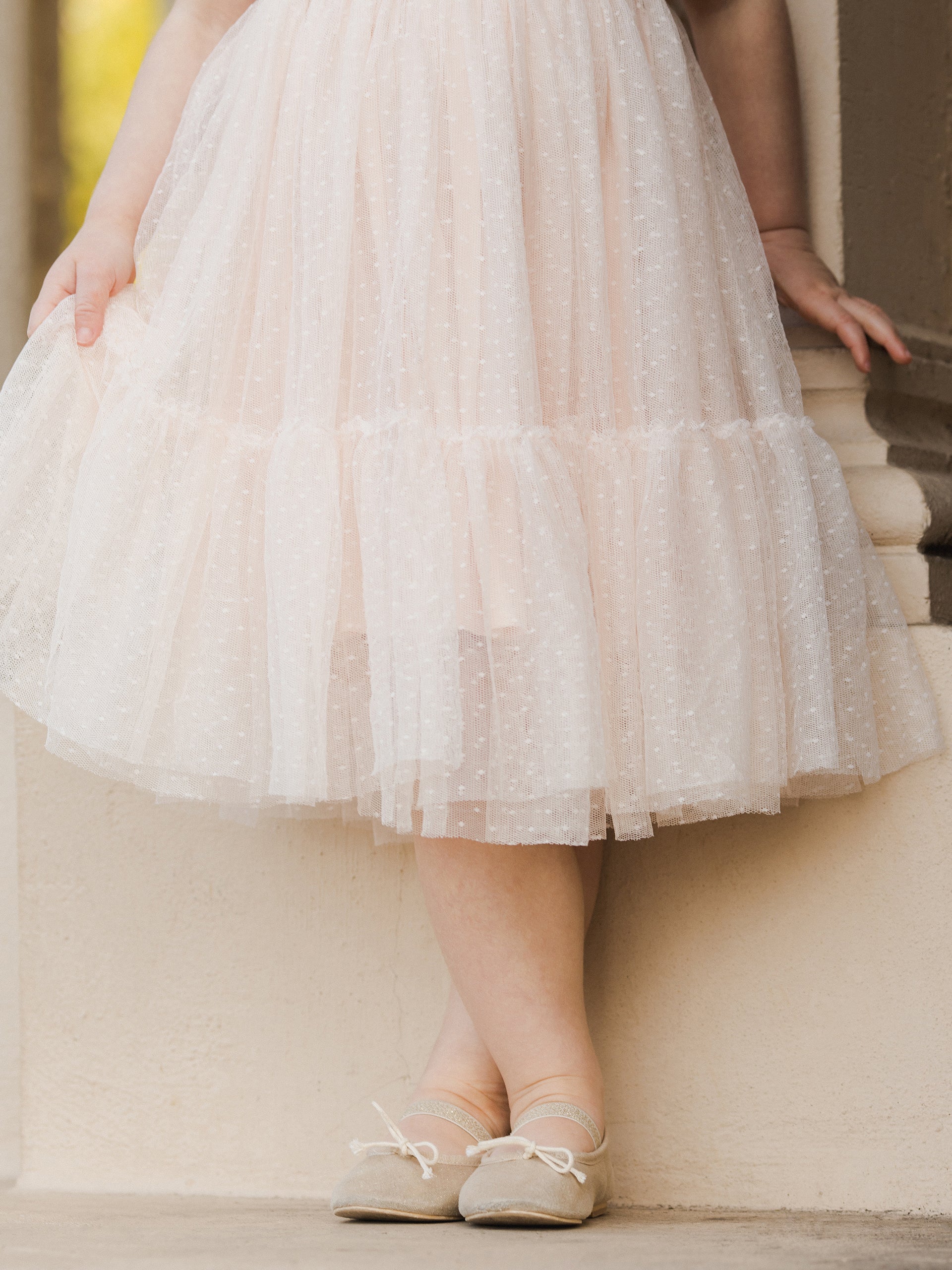 Child's feet in ballet flats, standing in a flowy pink skirt with layered tulle, against a light background.