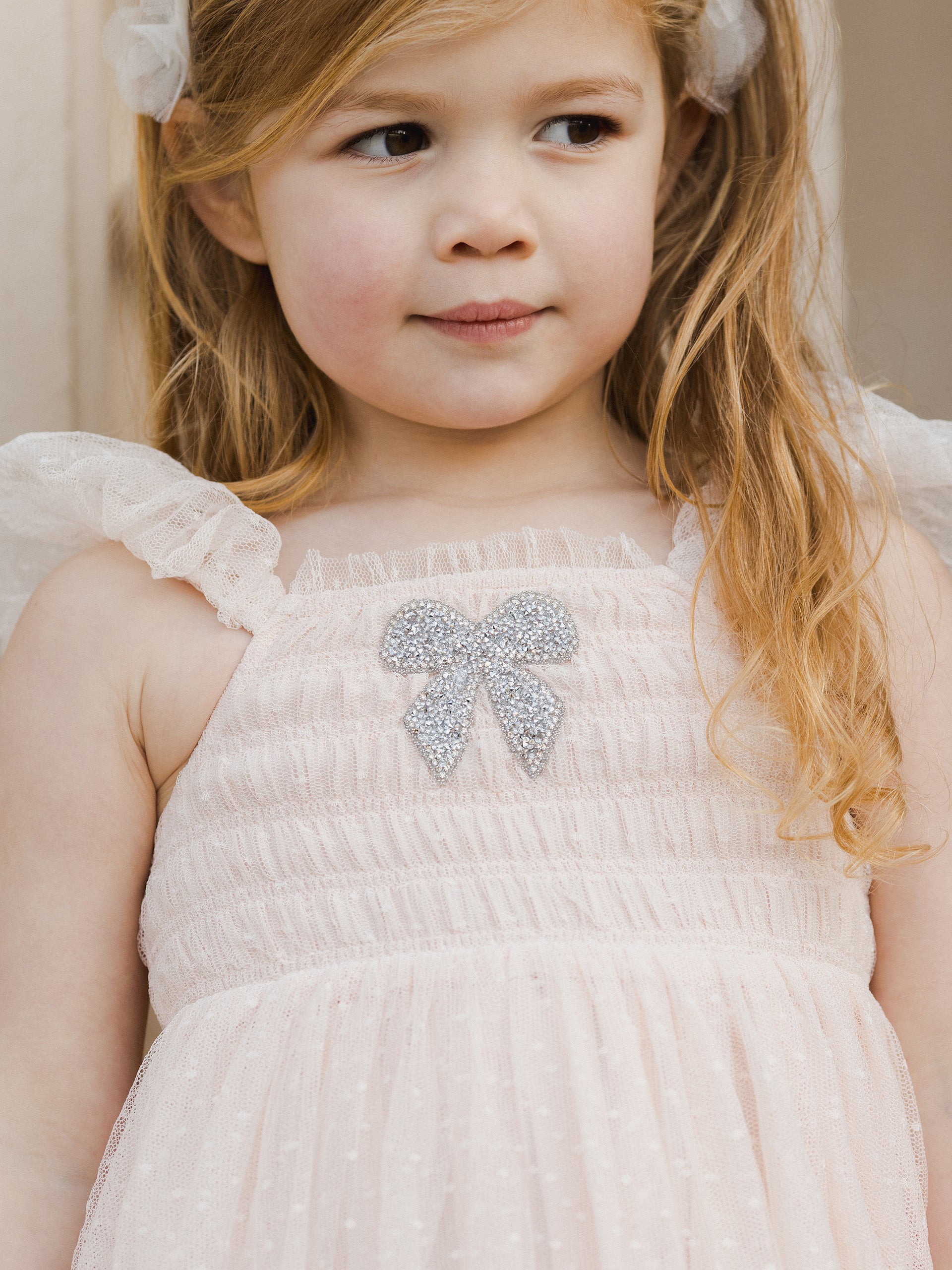 Girl with wavy hair in a pink dress featuring a glittery bow, looking thoughtfully at the camera.