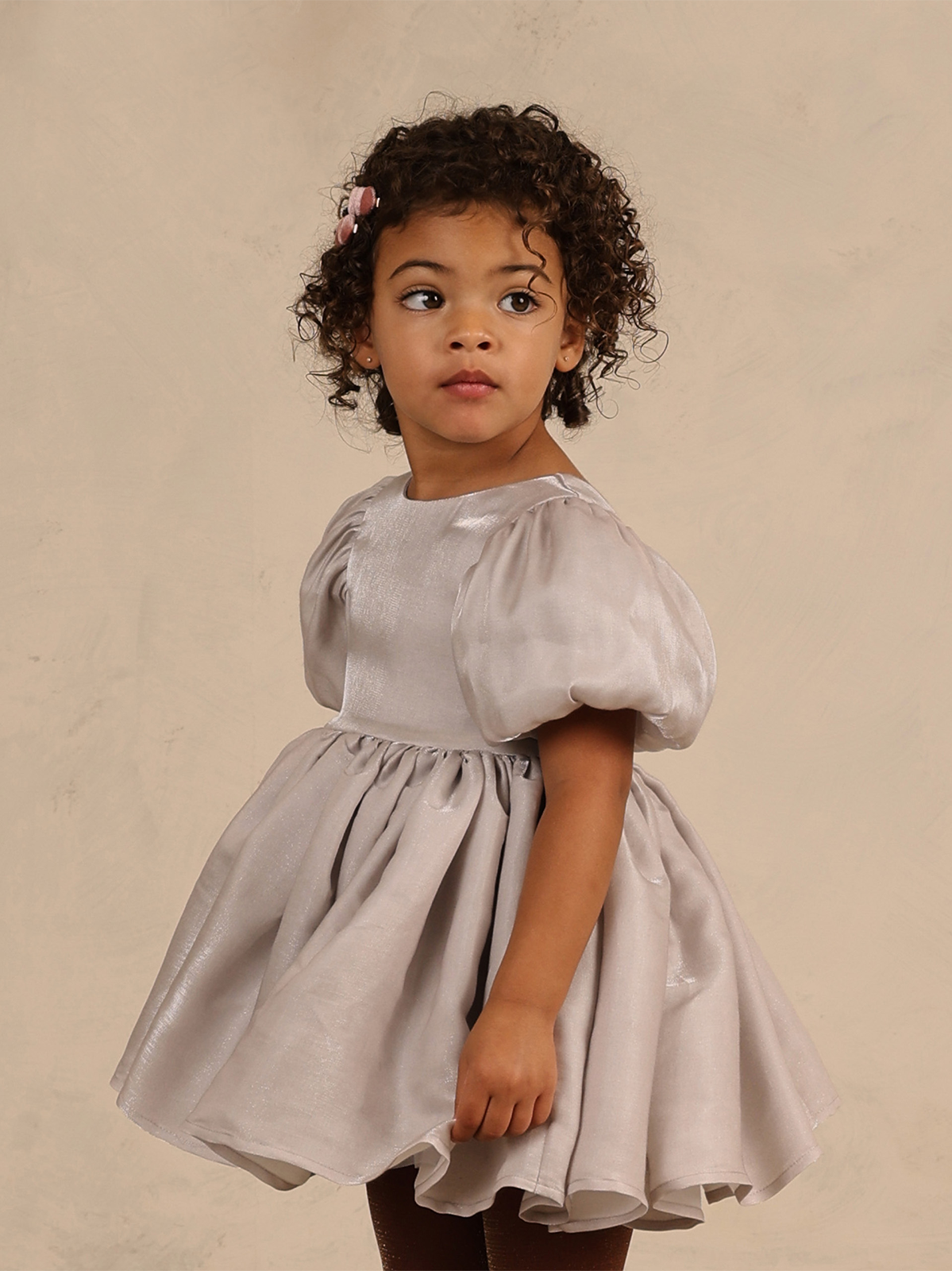 Child in a light gray dress, looking back over her shoulder with curly hair and a pink hair clip.