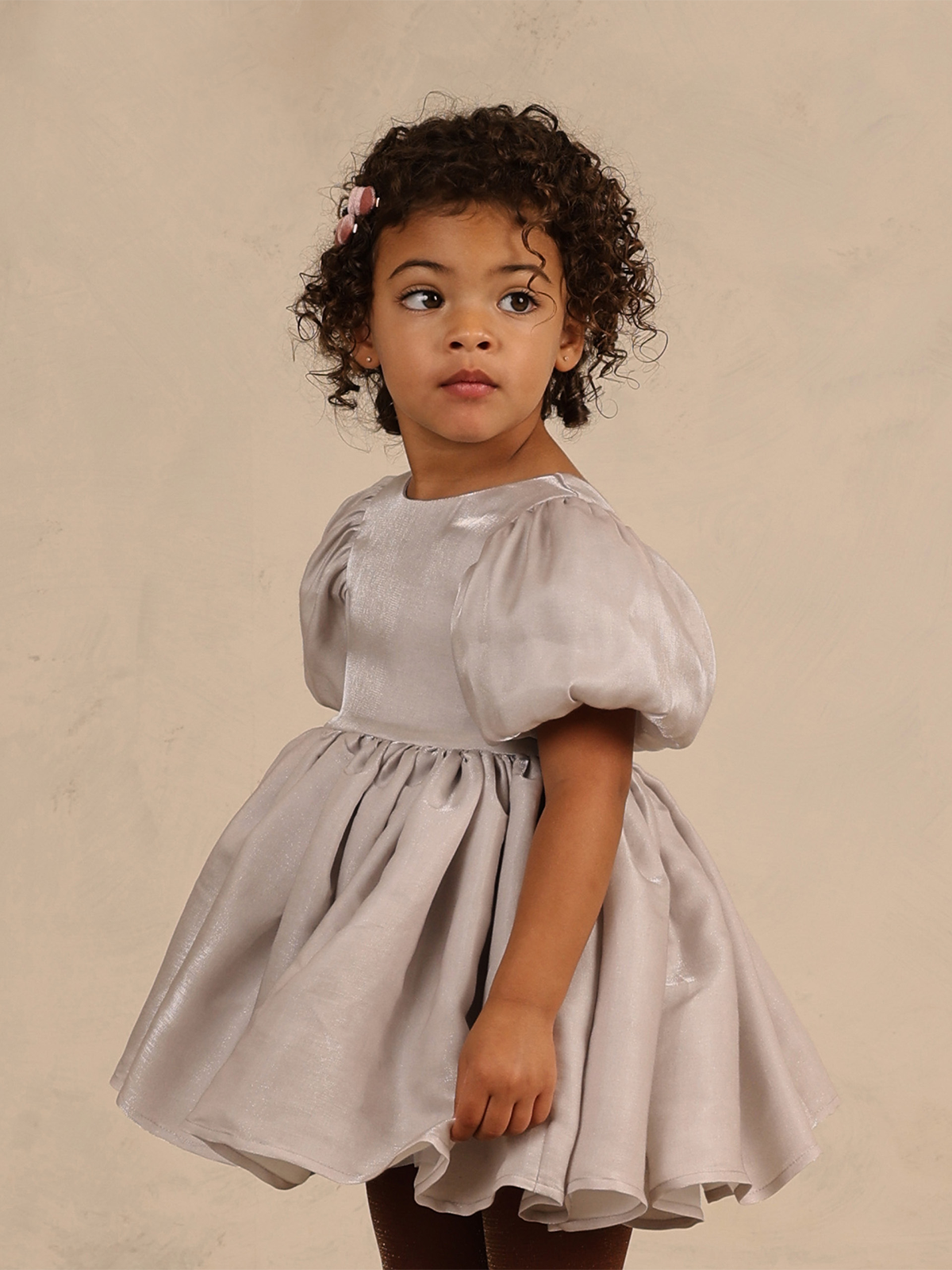 Child in a light gray dress, looking back over her shoulder with curly hair and a pink hair clip.
