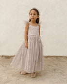 Girl in a light dress with fluffy shoulders stands on sandy ground against a textured white wall.