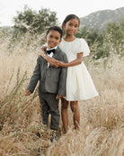 Two children happily pose together in a field, dressed in a suit and a white dress.
