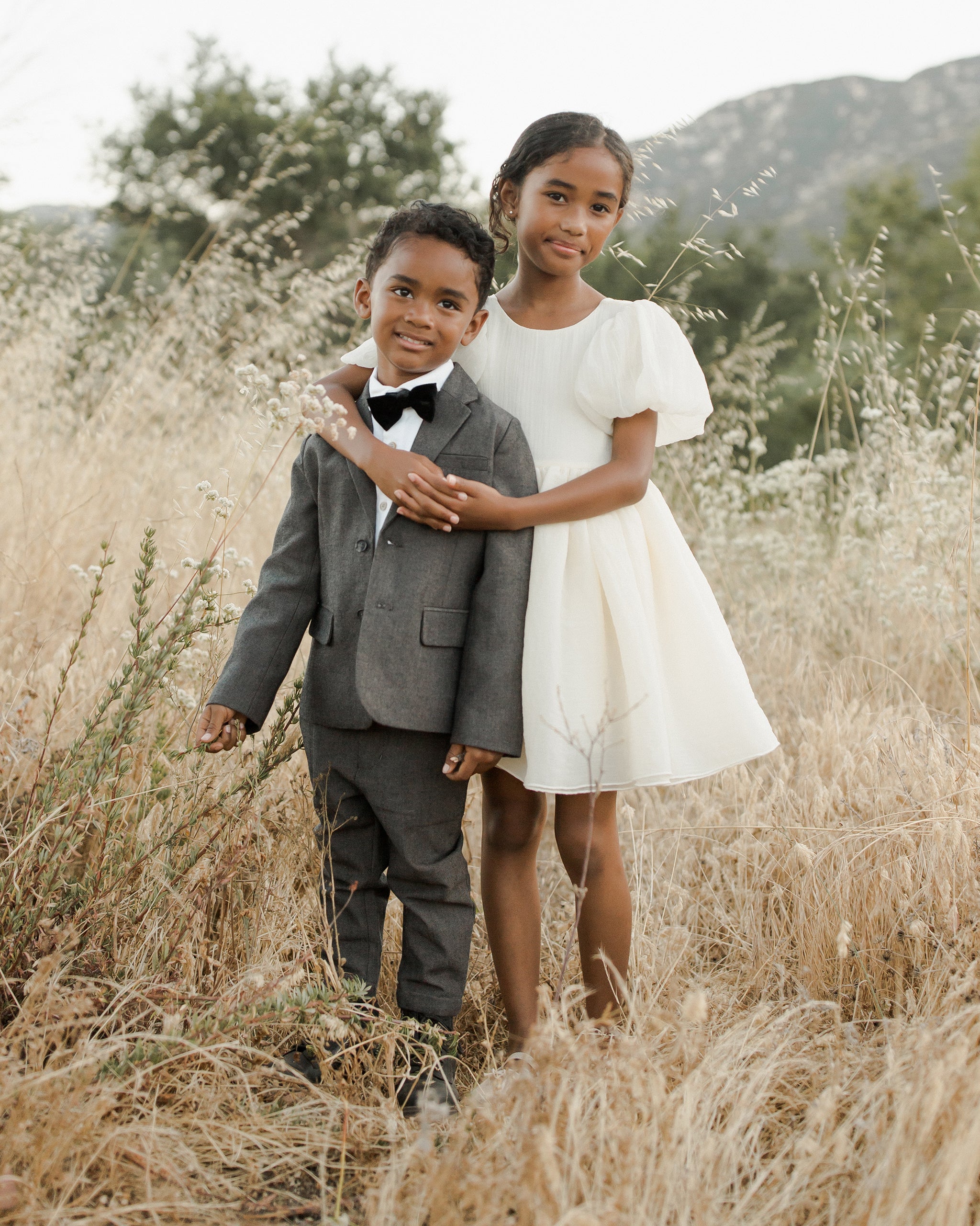 Two children happily pose together in a field, dressed in a suit and a white dress.
