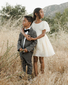Two children stand together in a field: a boy in a suit and a girl in a white dress, both looking away.