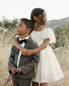 Two children stand in a field: a boy in a suit with a bow tie and a girl in a white dress, both smiling gently.