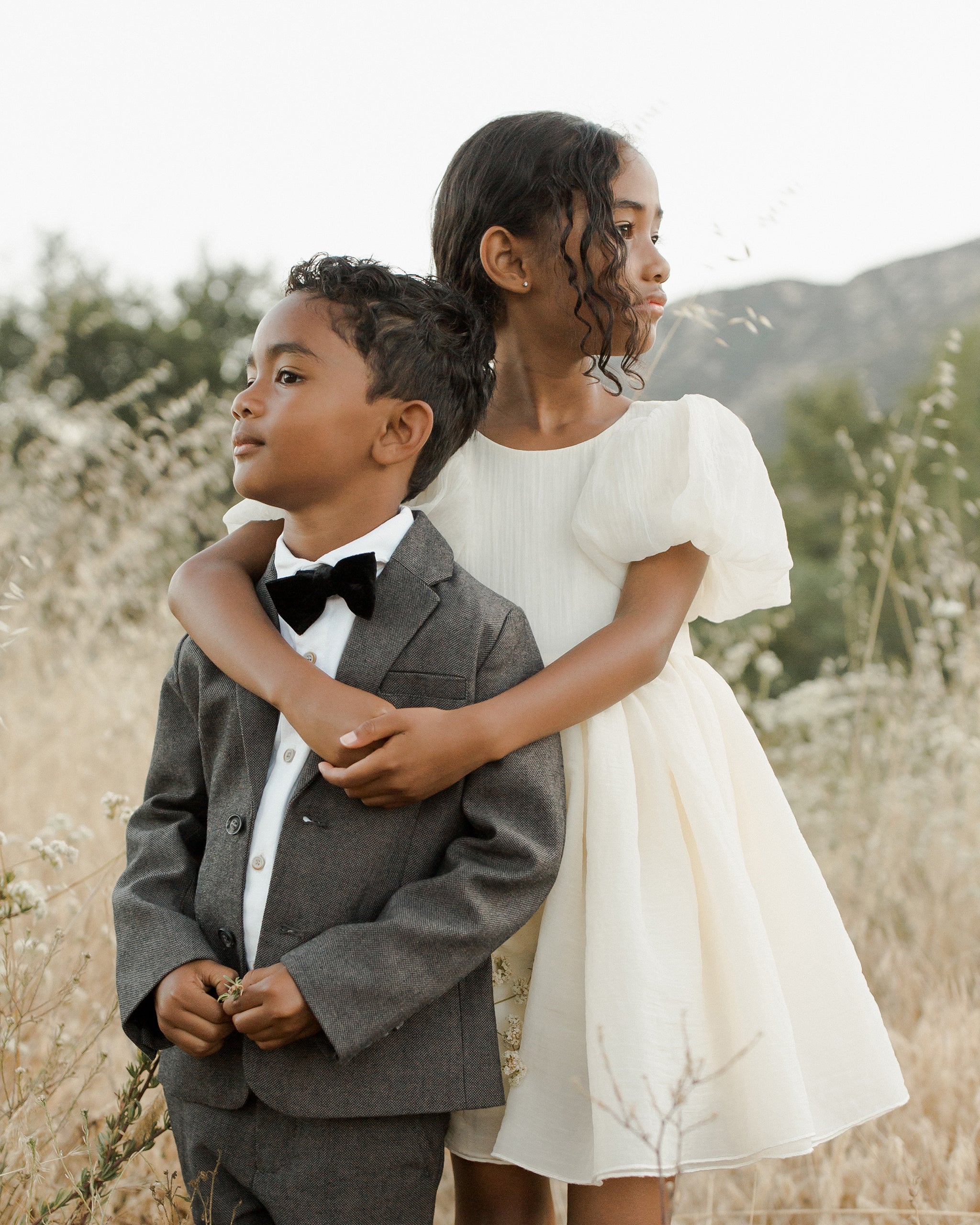 Two children stand in a field: a boy in a suit with a bow tie and a girl in a white dress, both smiling gently.