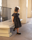Smiling child in black polka dot dress and bow, standing beside a stone bench in a well-lit area.