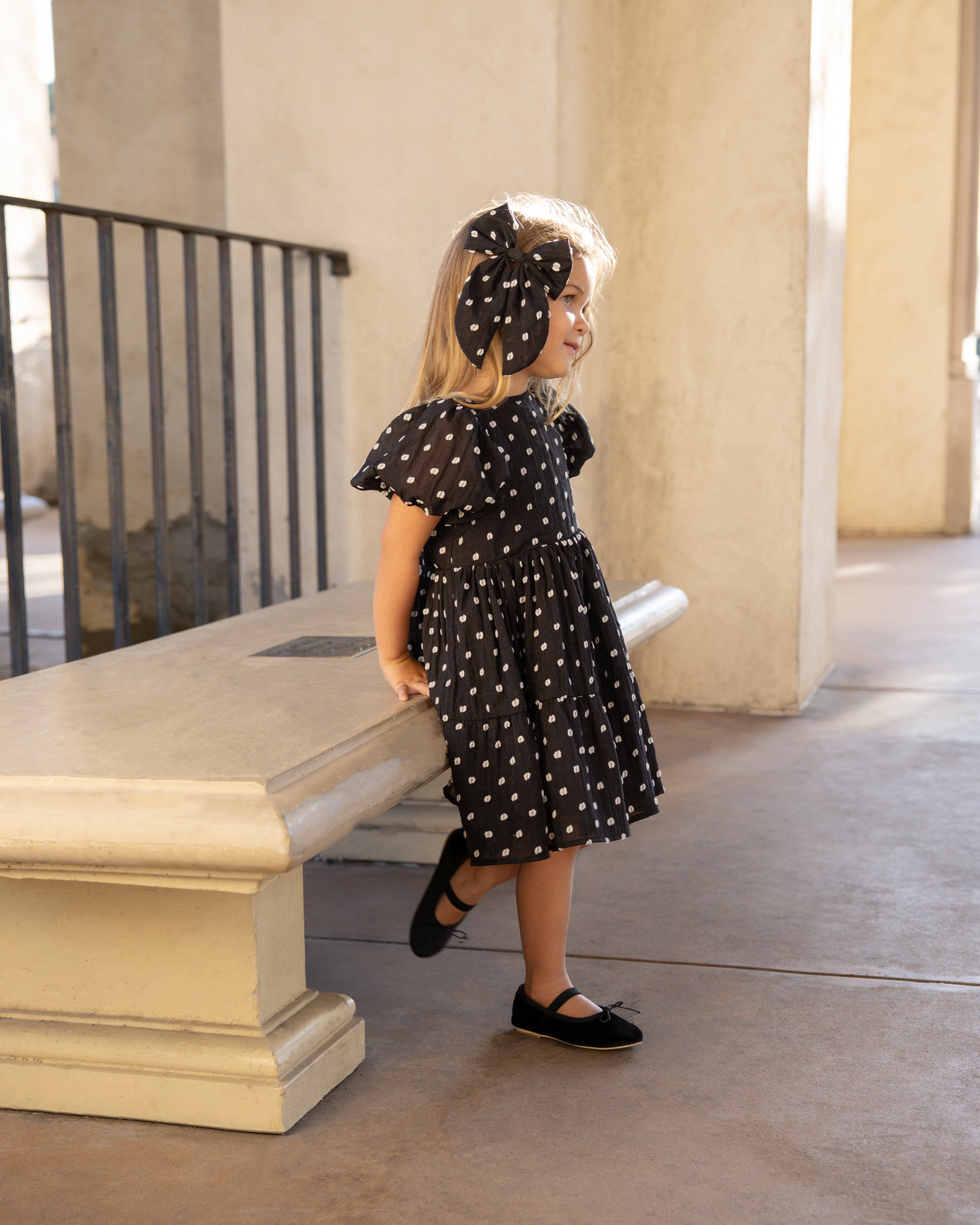 Smiling child in black polka dot dress and bow, standing beside a stone bench in a well-lit area.