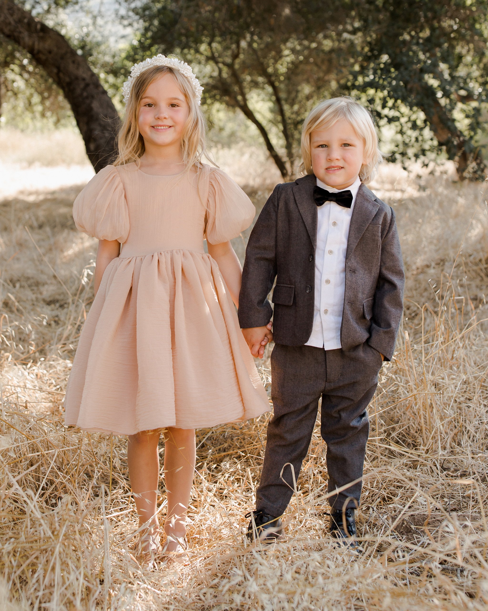 Two children hold hands in a field; a girl in a pink dress and a boy in a suit with a bow tie.