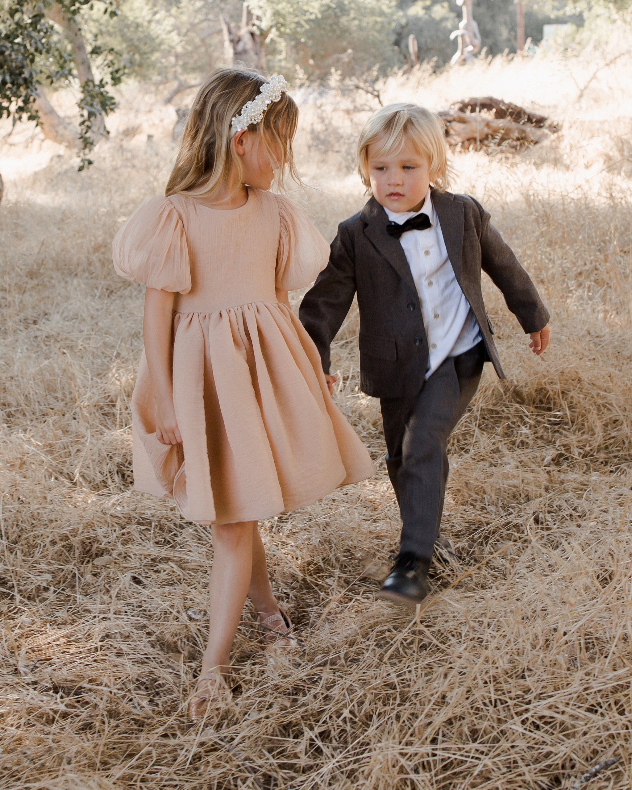 Two children walking in a field; a girl in a pink dress and a boy in a suit with a bow tie.