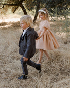 Two children walking hand in hand in a grassy field, dressed in formal attire.