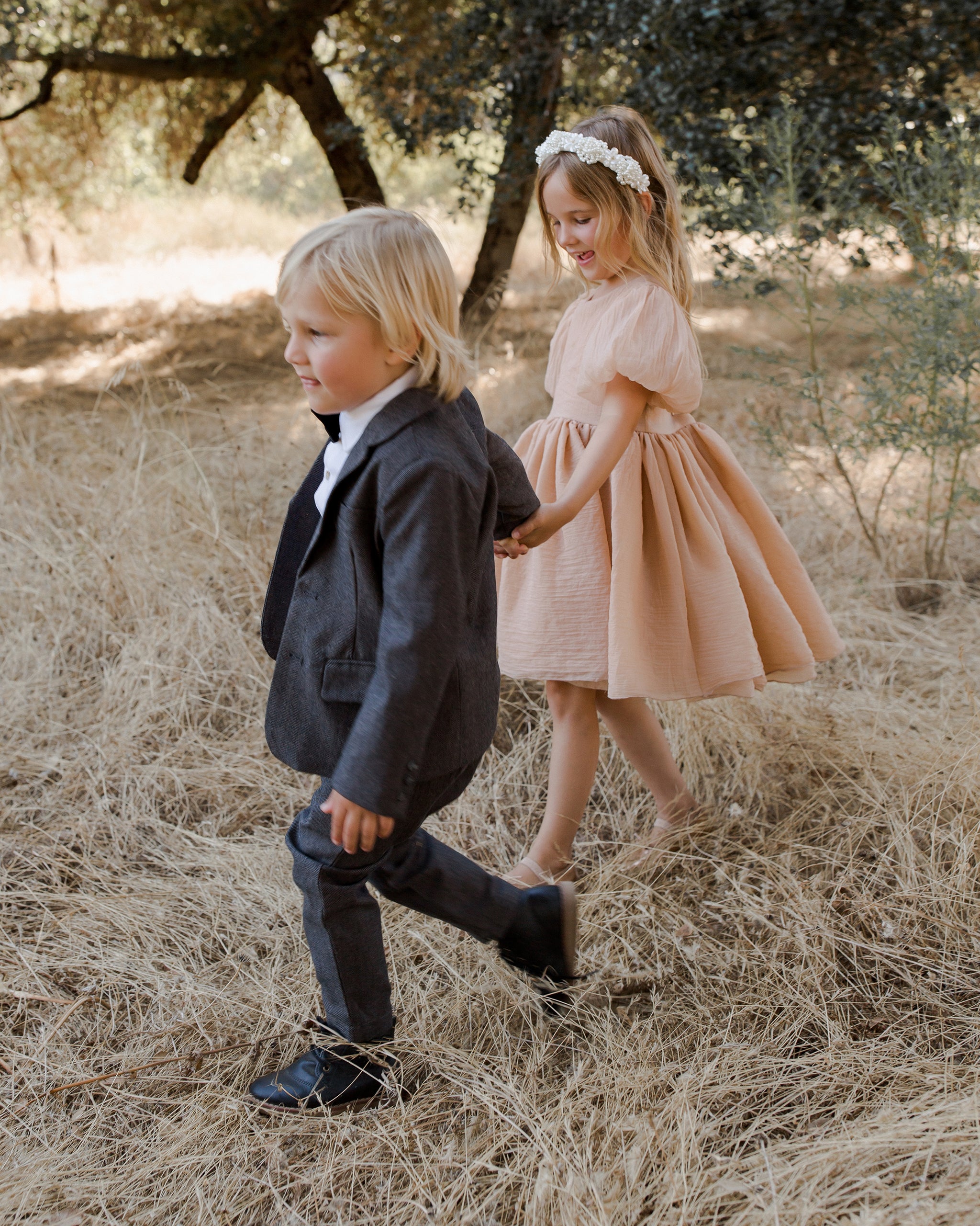 Two children walking hand in hand in a grassy field, dressed in formal attire.