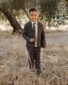 A smiling boy in a gray suit stands in dried grass near trees, looking confidently at the camera.