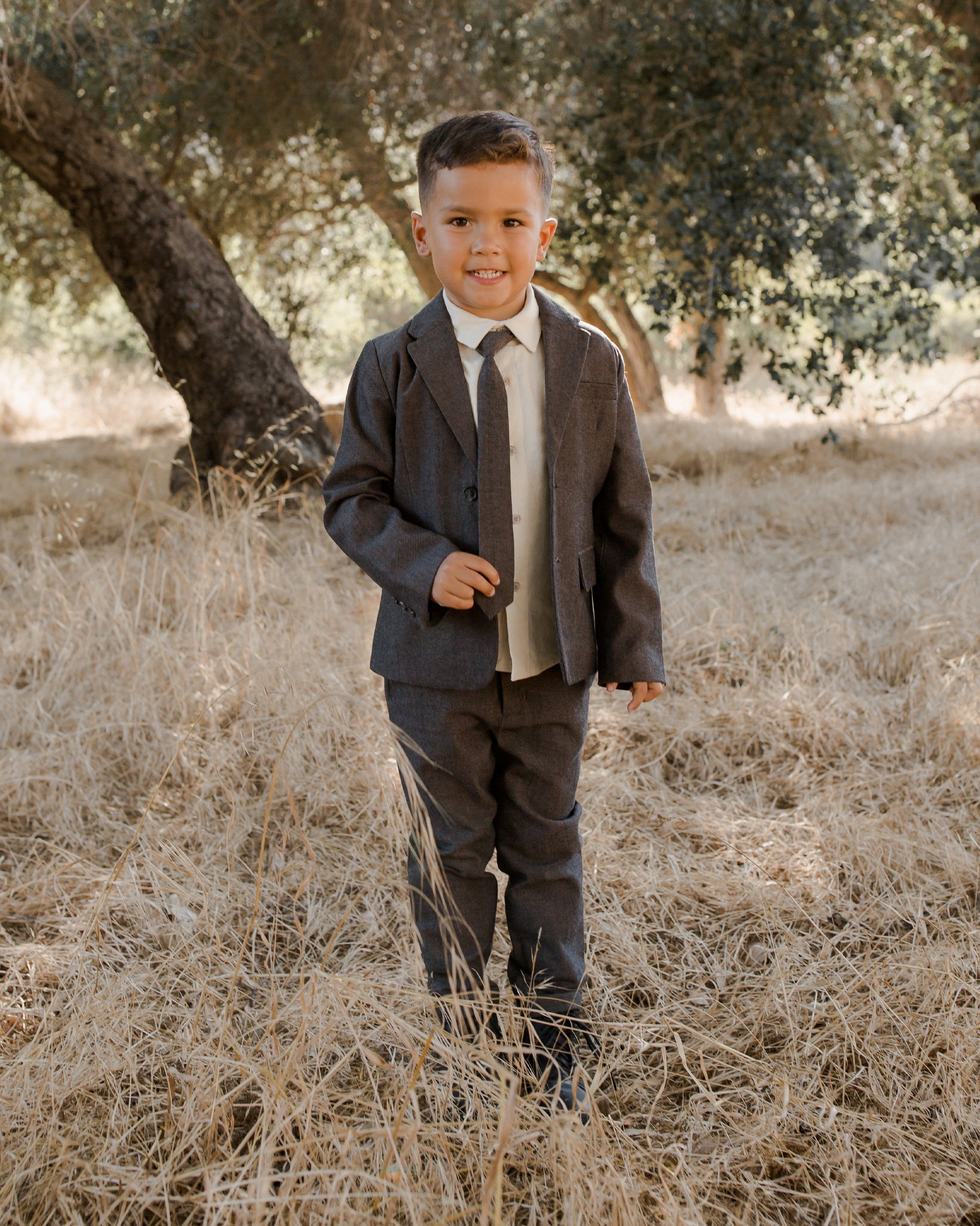 A smiling boy in a gray suit stands in dried grass near trees, looking confidently at the camera.