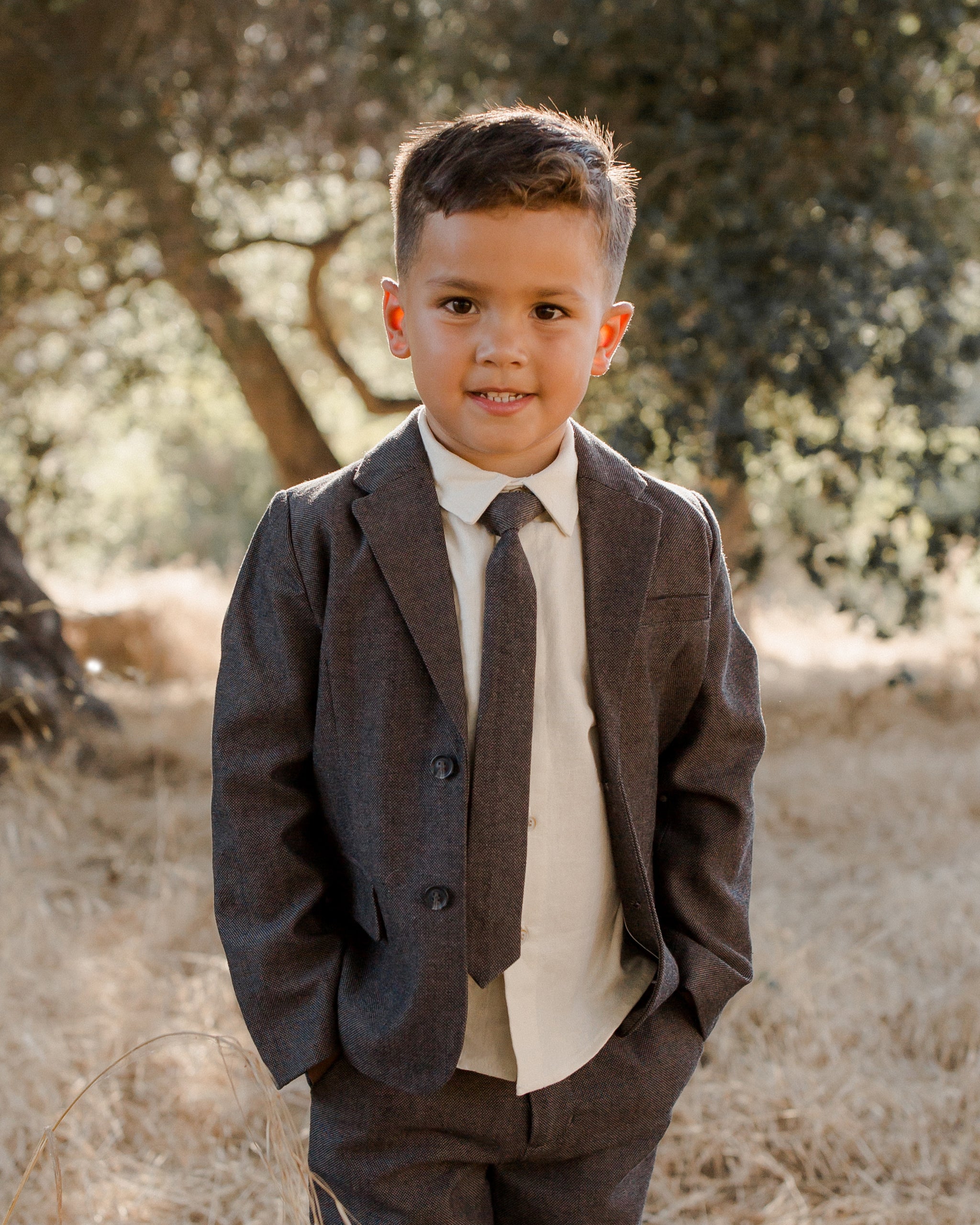 Young boy in a suit smiles confidently in a sunny, outdoor location with trees and dry grass.