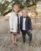 Two children in formal attire pose in a field of tall grass, surrounded by trees.