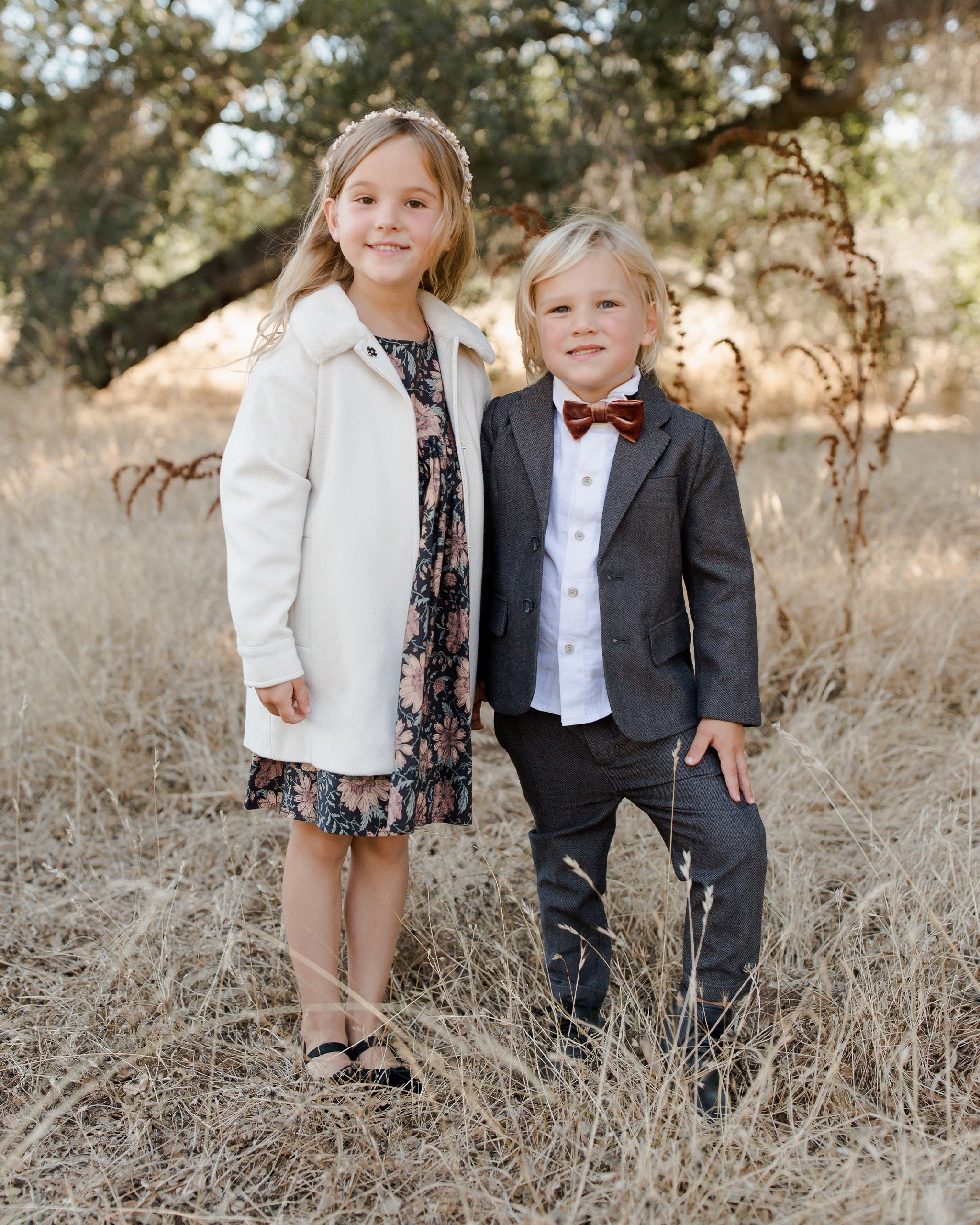 Two children in formal attire pose in a field of tall grass, surrounded by trees.