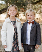 Two smiling children: a girl in a floral dress and a boy in a suit with a bow tie, standing outdoors.