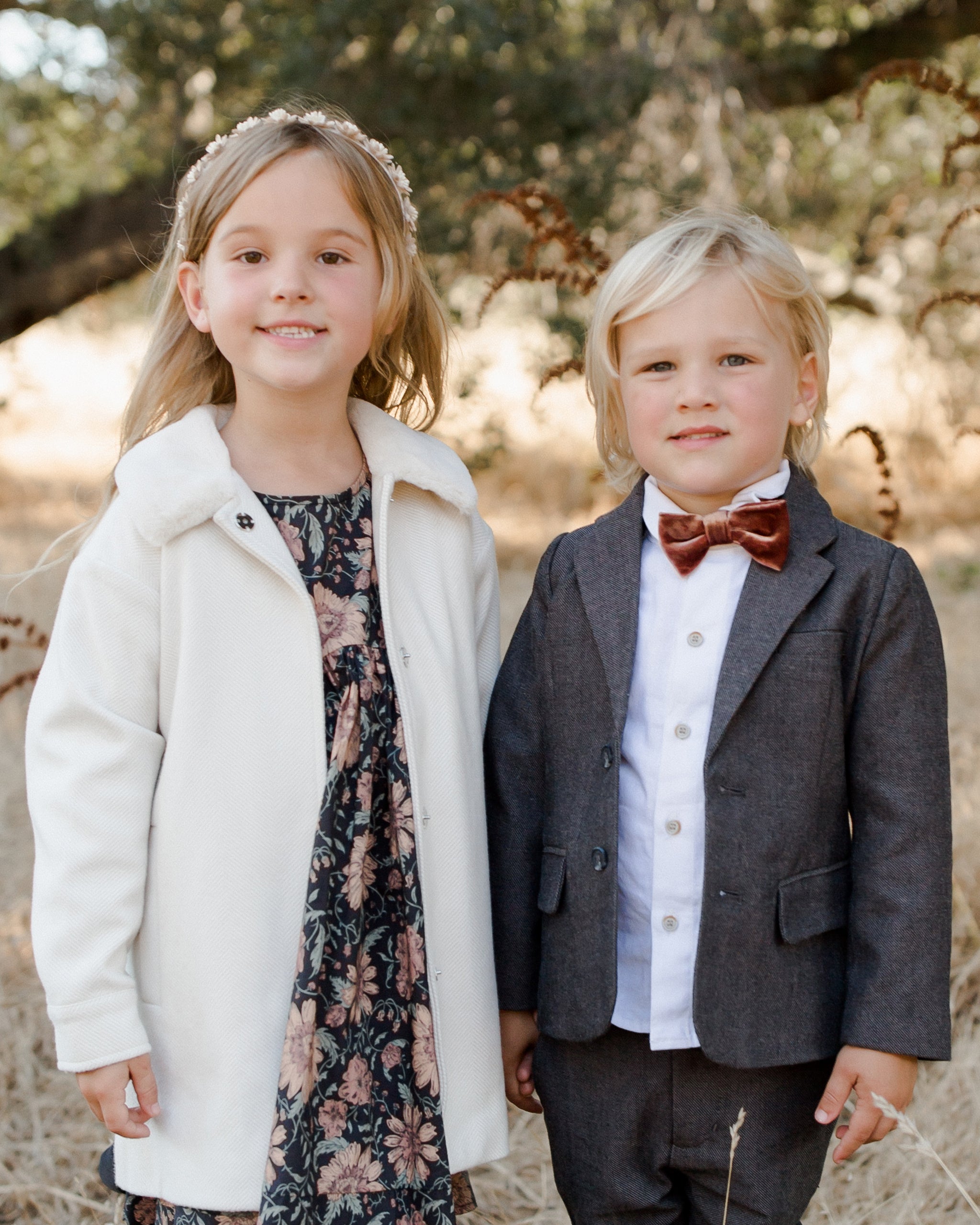 Two smiling children: a girl in a floral dress and a boy in a suit with a bow tie, standing outdoors.