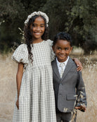 Two children stand together in a field, smiling; the girl wears a checkered dress and headband, the boy in a suit.