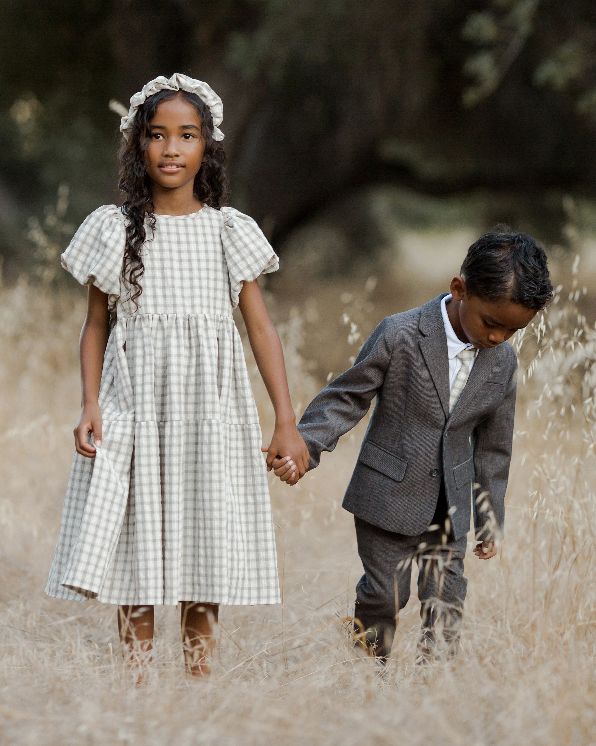 Two children hold hands in a field, wearing a dress and suit, surrounded by tall golden grass.
