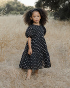 Young girl with curly hair wearing a polka dot dress, standing in a field of tall grass.