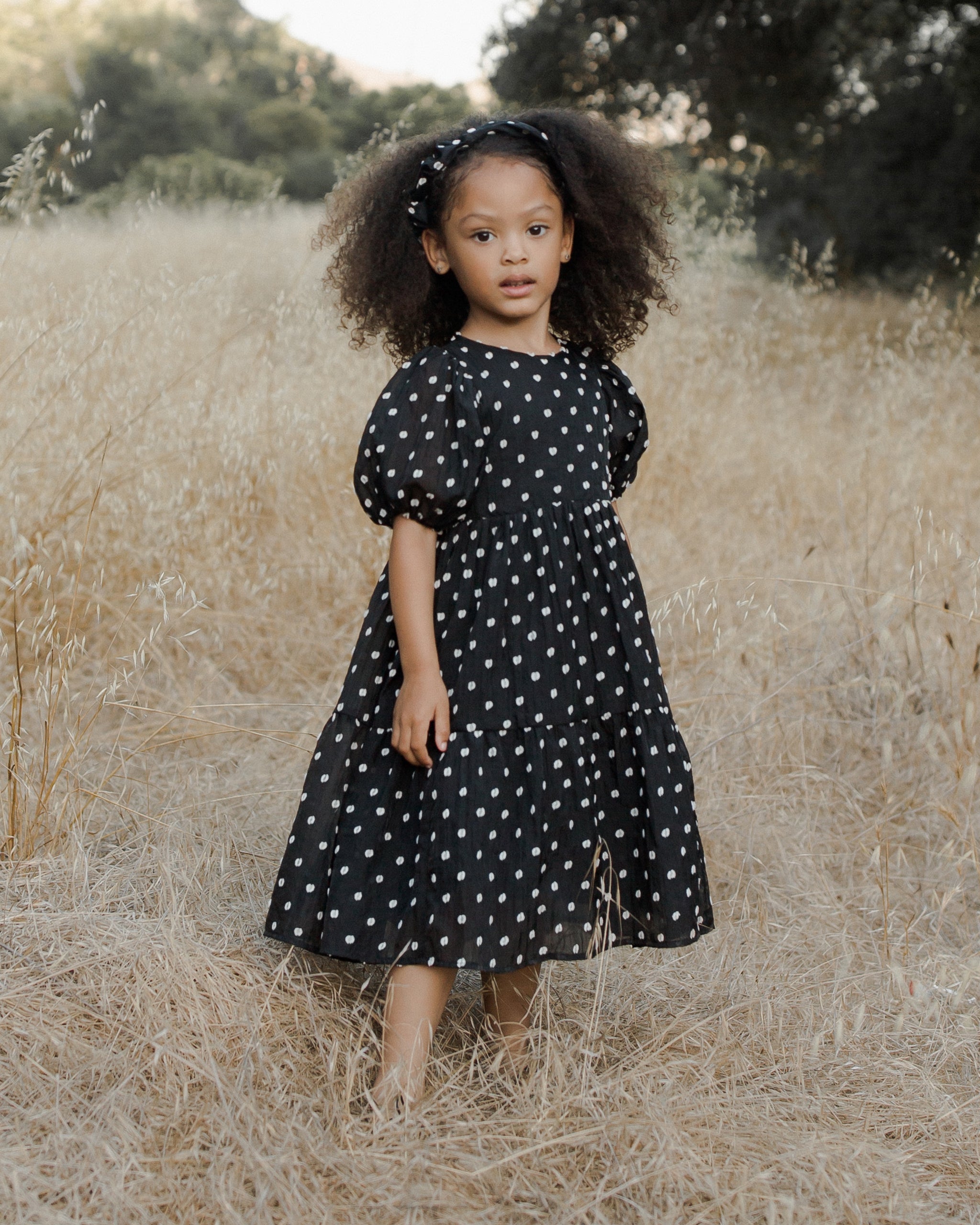 Young girl with curly hair wearing a polka dot dress, standing in a field of tall grass.