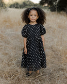 Girl with curly hair in a black polka dot dress standing in a dry field, smiling at the camera.