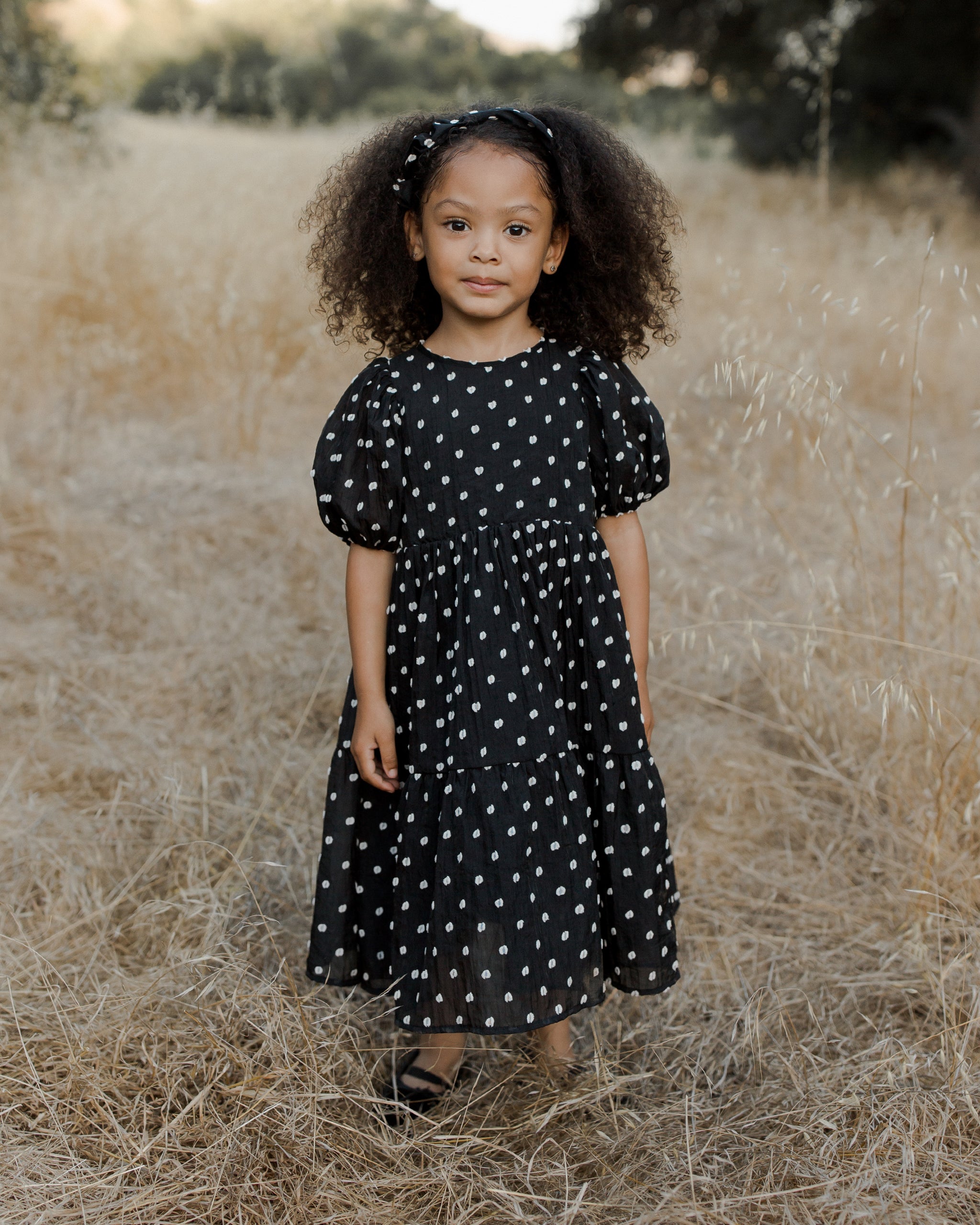Girl with curly hair in a black polka dot dress standing in a dry field, smiling at the camera.