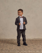 Smiling young boy in a gray suit and bow tie, standing on a patterned rug against a neutral background.