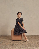 Girl in a black polka dot dress and ballet shoes sitting on a stool, smiling brightly.