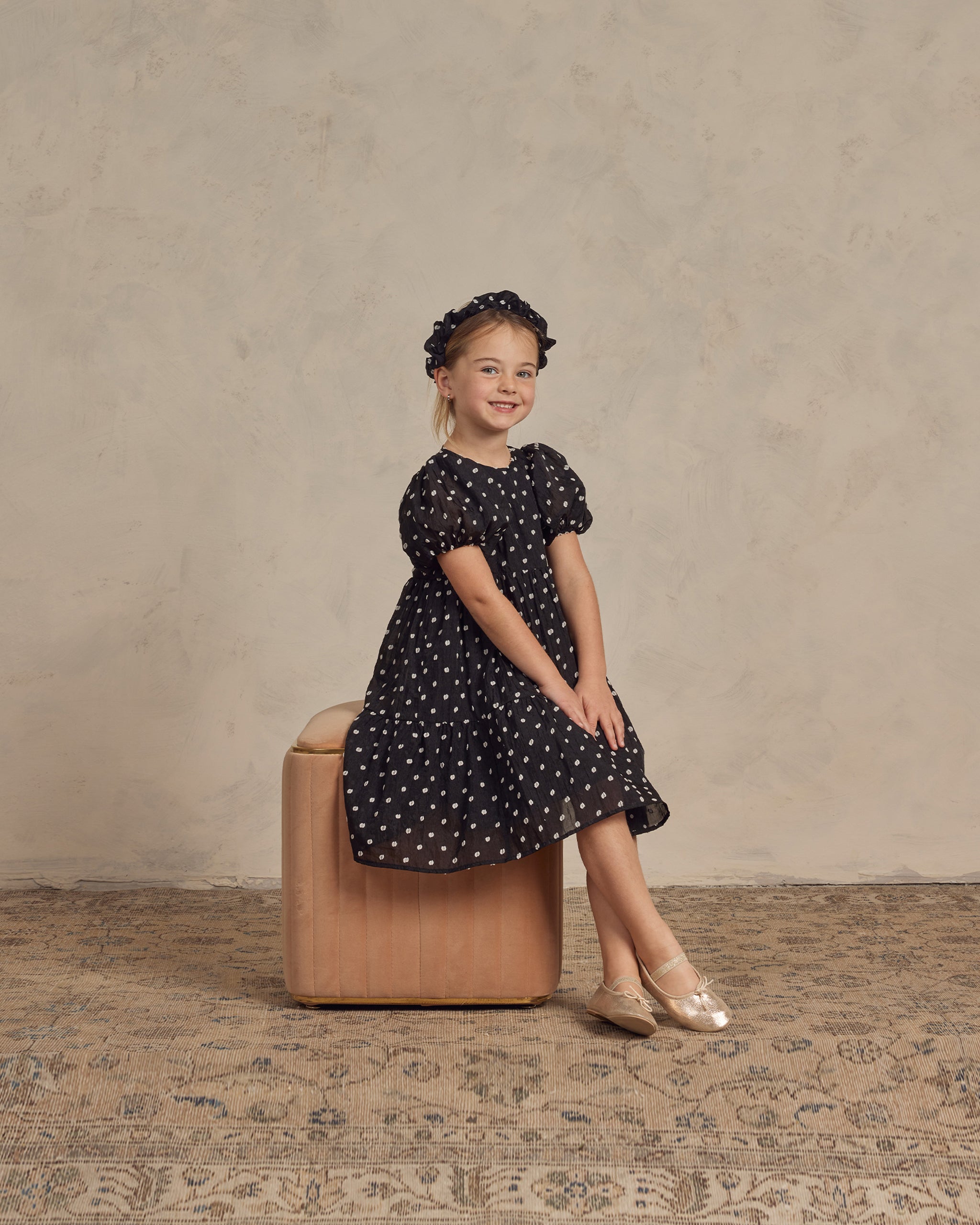 Girl in a black polka dot dress and ballet shoes sitting on a stool, smiling brightly.