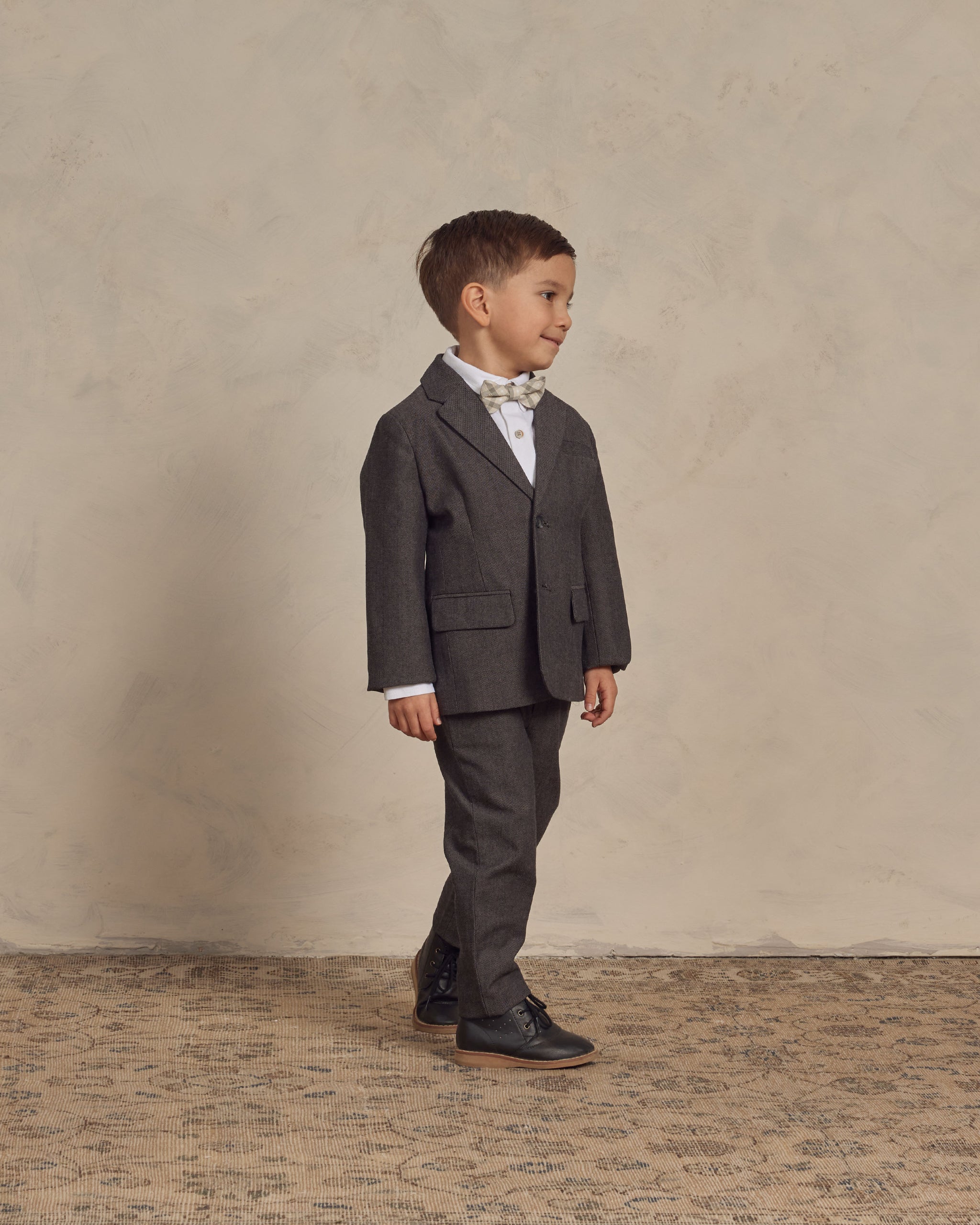 Boy in a gray suit with a bow tie, standing on a patterned rug against a textured background.