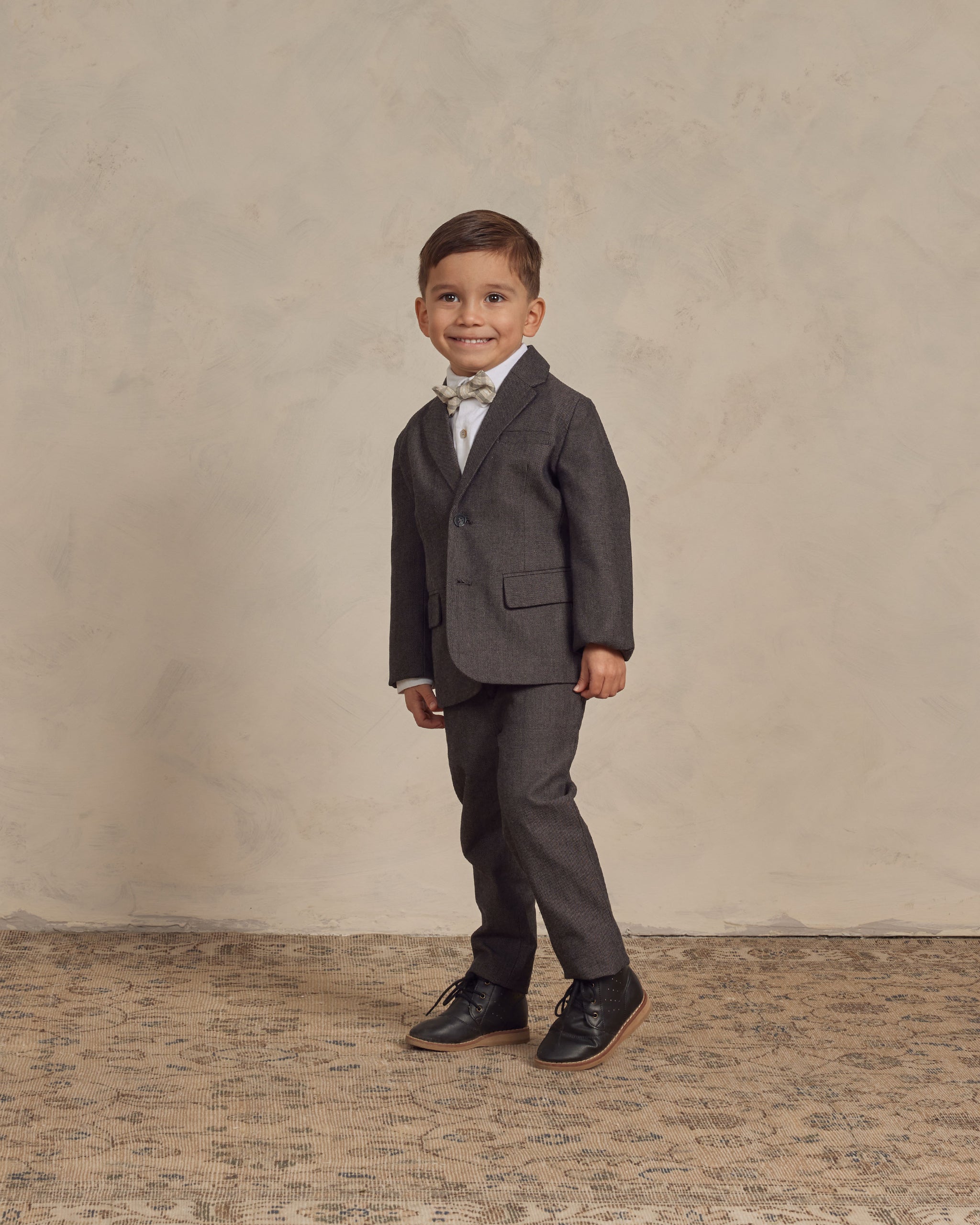 A smiling boy in a dark suit and bow tie poses confidently on a textured floor against a neutral backdrop.