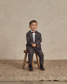 Smiling boy in a gray suit and bow tie sitting on a wooden stool against a textured backdrop.