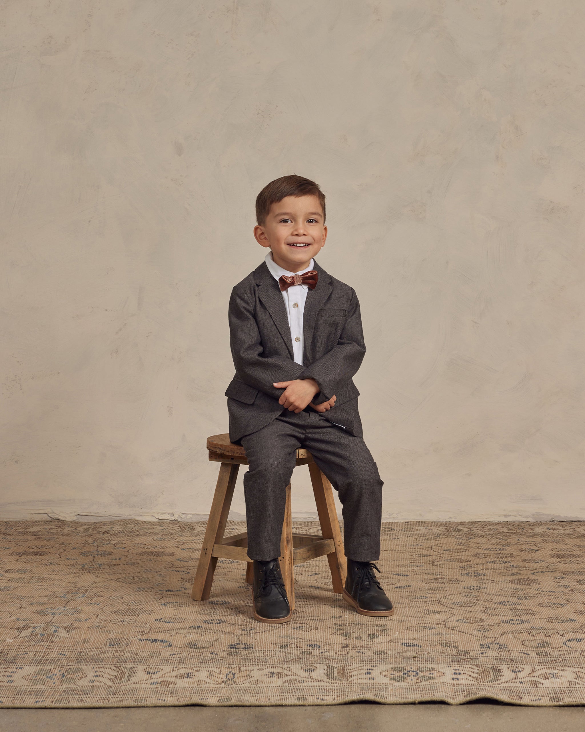 Smiling boy in a gray suit and bow tie sitting on a wooden stool against a textured backdrop.
