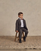 Boy in a formal gray suit with a bow tie sitting on a wooden stool against a neutral background.