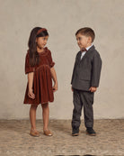 Two children interact: a girl in a rust dress and a boy in a gray suit, smiling against a textured background.
