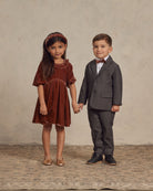 Two children standing together, the girl in a brown dress and the boy in a grey suit holding hands.