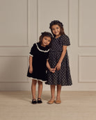 Two young girls standing together, one in a black velvet dress and the other in a polka dot dress, smiling.