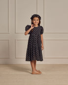 Girl in a black polka dot dress and matching headband, smiling and standing in a minimalist room.