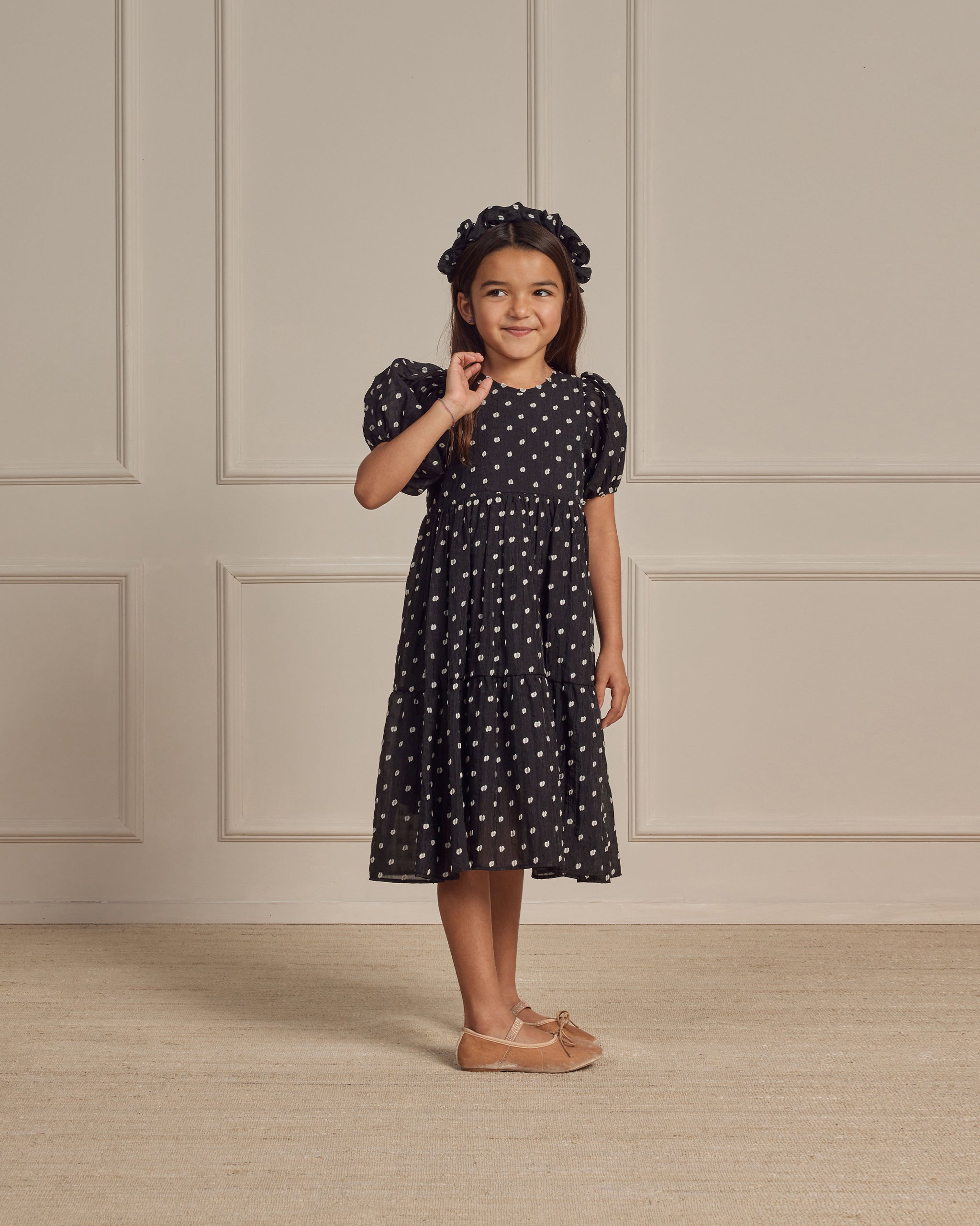 Girl in a black polka dot dress and matching headband, smiling and standing in a minimalist room.